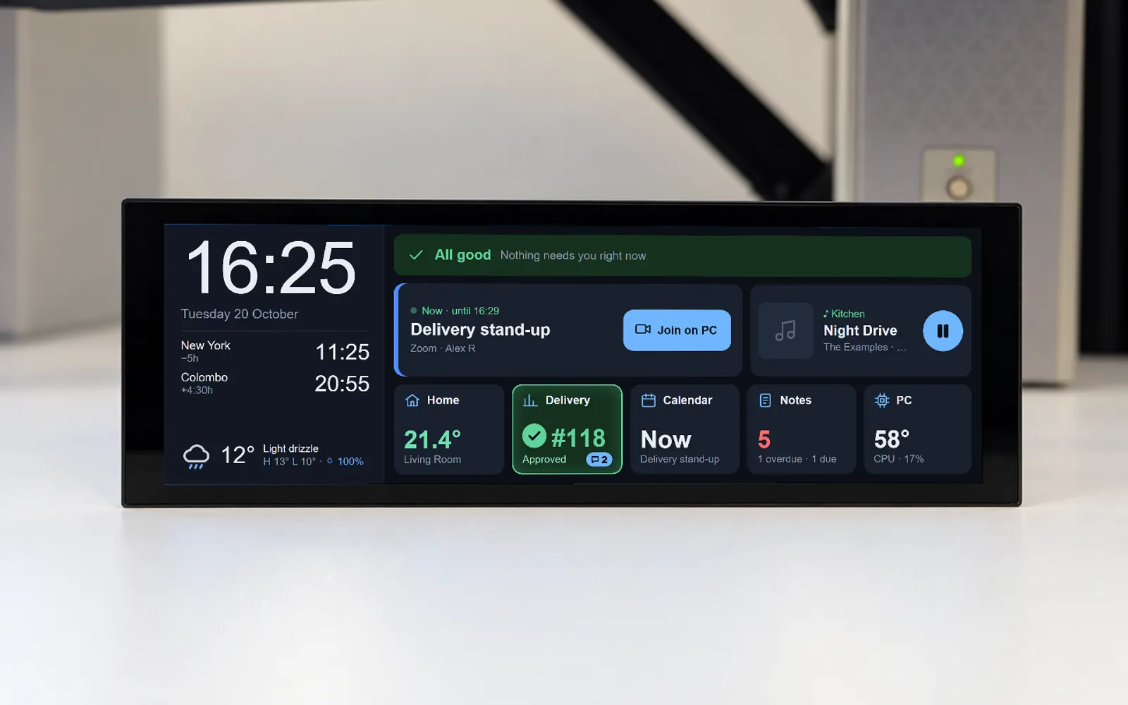 A render of the Pi Dash unit standing on a desk below two monitors — a wide, thin black touch panel showing a clock, two world clocks and the weather on the left, and an "All good" strip, the next meeting with a "Join on PC" button, a now-playing card and five status tiles on the right